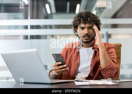 Junger Mann erlebt Stress und Verwirrung beim Arbeiten an einem Laptop und hält ein Smartphone, berührt mit gerunzelter Stirn seine Schläfe in einem modernen o Stockfoto