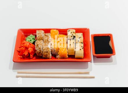 Frische verschiedene Sushi-Rollen, serviert auf einem lebhaften roten Teller mit Sojasauce, Wasabi, Ingwer und Stäbchen vor einem sauberen weißen Hintergrund. Traditionelle japanische Küche, farbenfroher Meeresfrüchte-Vorspeise, gesundes asiatisches Lebensmittelkonzept mit Platz für Text. Stockfoto