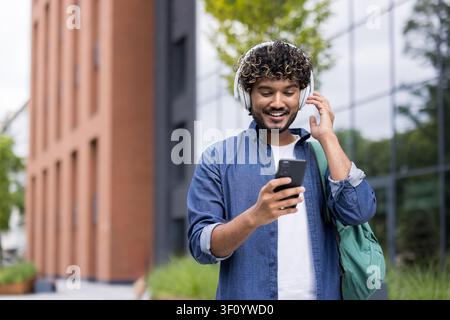 Lächelnder indischer Student trägt Kopfhörer und einen Rucksack, geht im Freien und schaut auf sein Smartphone, genießt Musik oder einen Podcast Stockfoto