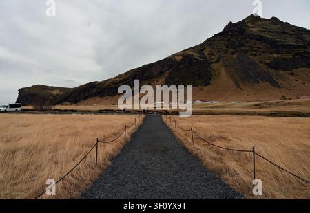 Schotterweg, gesäumt von Kettenzäunen, schneidet sich durch goldene Winterwiese in Richtung eines gezackten Vulkanbergs Stockfoto