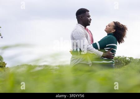 Afroamerikanisches Paar umarmt sich auf einer Wiese, trägt gestreiften Pullover und rosa T-Shirt, Kopierraum Stockfoto