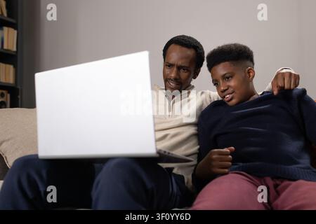 Afroamerikanischer Vater und Teenager-Sohn sitzen im Wohnzimmer auf dem Sofa und schauen gemeinsam auf den Laptop. Familie, Zuhause, Couch, Computer, Bücherregal, gemütlich, bo Stockfoto