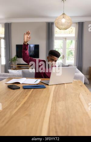 Teenager afrikanisch-amerikanischer Herkunft in burgunderrotem Pullover, der am Heimtisch mit einem silbernen Laptop lernt und Notizen macht Stockfoto