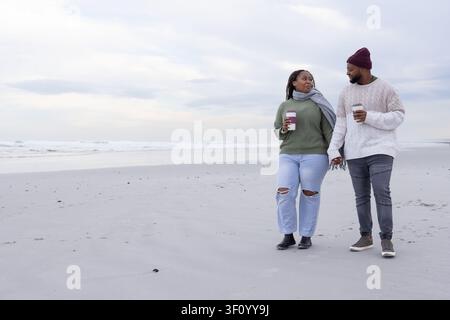 Paar geht an breitem Sandstrand entlang, hält sich an den Händen und trägt To-go-Becher, mit Beanie und Schal, Kopierraum Stockfoto