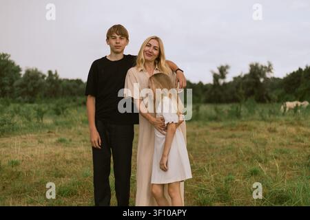 Eine Mutter umarmt liebevoll ihren Sohn und ihre Tochter, während sie auf einem weiten grünen Feld steht, mit Pferden, die friedlich in der Ferne grasen, unter einer bewölkten Stockfoto