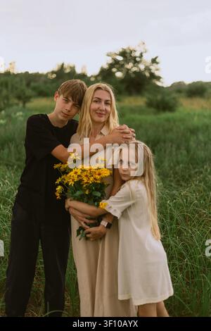 Eine liebevolle Mutter hält ihre Kinder eng an sich gedrückt, während sie auf einem üppigen grünen Feld steht, umgeben von der Schönheit der Natur und einem farbenfrohen Blumenarrangement Stockfoto