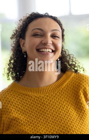Frau lächelt, neigt den Kopf, trägt einen Senfgelb-Pullover und goldene Kreolen mit Perlen in der Nähe eines großen Fensters Stockfoto