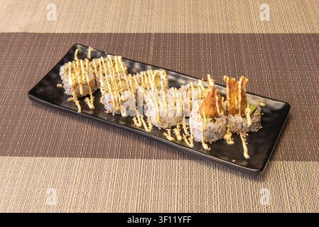 Verschiedene Sushi-Rollen mit Tempura-Garnelen, Sesamsamen und cremiger Sauce, die auf einem schwarzen Teller geträufelt ist Stockfoto