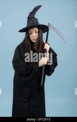 Ernsthafte junge Frau in einem Hexenkostüm mit einer großen Sense Stockfoto