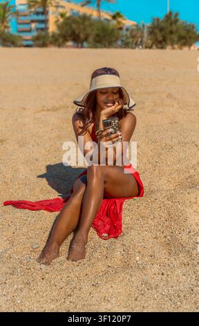 Lächelnde junge kubanische Frau in rotem Bikini und Sonnenhut, die auf einem Sandstrand sitzt und ein Mobiltelefon benutzt Stockfoto