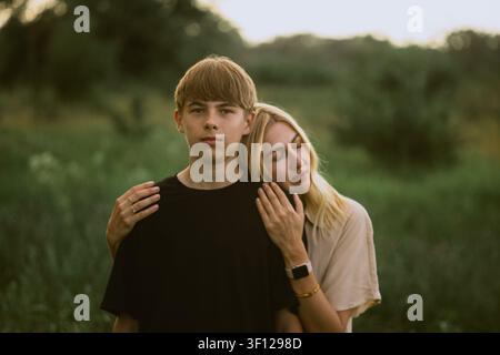 Ein junger Mann und eine Frau teilen einen Moment der Zuneigung im Freien, wobei die Frau ihren Kopf auf seiner Schulter ruht, umgeben von üppiger Grünflora Stockfoto