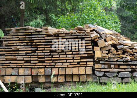 Raue Schnittholzplanken und schwere Holzbauteile sind im Freien in der Nähe von grünem Gras und Waldbäumen gestapelt und stellen Rohmaterialien für den Bau bereit Stockfoto