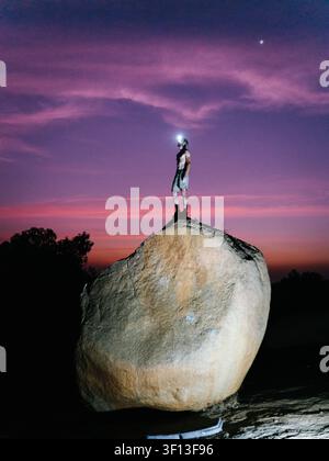 Ansicht eines Mannes mit einer Stirnlampe, der während der Dämmerung auf einem großen Granitfelsen in Hampi, Karnataka, Indien steht. Stockfoto