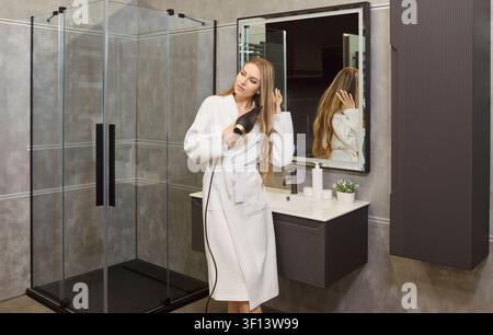Attraktive Frau in einem modernen Badezimmer, trägt einen weißen Bademantel und steht, während sie ihre Haare föhnt und stylt Stockfoto