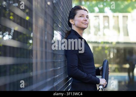 Frau Mitte 40 in Sportkleidung auf dem Platz, lehnt sich an einen Drahtzaun, hält ein Paddel und blickt nach rechts, Kopierraum Stockfoto