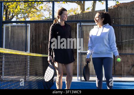 Verschiedene Freundinnen lächeln in Sportkleidung und halten Schläger mit Ball auf blauem Platz nahe dem Netz Stockfoto