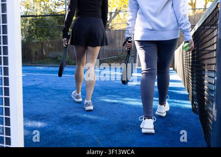 Verschiedene Freundinnen gehen auf einem blauen Paddelplatz in Sportkleidung und tragen Paddel und grünen Ball Stockfoto