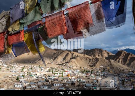 Bunte ausgefranste tibetische Gebetsfahnen wehen im Vordergrund und öffnen sich zu einem weiten panoramischen Hochwinkelblick über die Stadt Leh und den majestätischen Barr Stockfoto