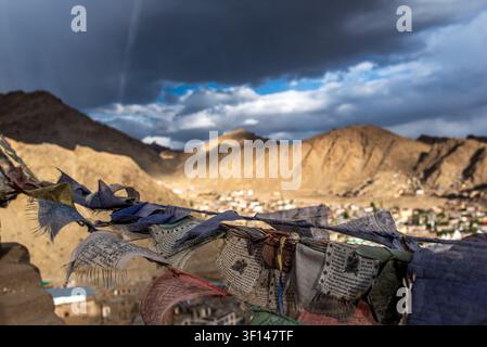 Bunte ausgefranste tibetische Gebetsfahnen wehen im Vordergrund und öffnen sich zu einem weiten panoramischen Hochwinkelblick über die Stadt Leh und den majestätischen Barr Stockfoto