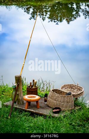 Eine rustikale, altmodische Angelausrüstung auf einem Holzsteg an einem ruhigen Teich, bestehend aus einer Bambus-Angelrute, gewebten Weidenkörben und einem kleinen Holzschemel. Stockfoto
