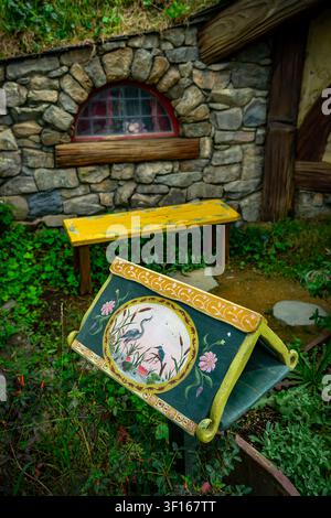Ein wunderschön handbemalter hölzerner Bücherständer mit traditionellen Blumen- und Vogelillustrationen steht vor einem steinernen Hobbit-Haus mit einer gelben Bank Stockfoto
