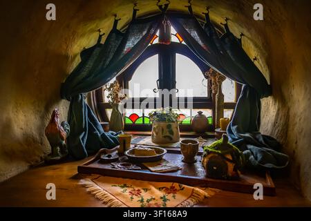 Ein intimer, gemütlicher Innenblick auf ein Fenster eines Bag-End-Hobbitlochs am Hobbiton-Filmset, mit einem rustikalen hölzernen Teetablett mit Kuchen und Keramikbechern. Stockfoto