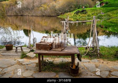 Eine rustikale hölzerne Fischereistation mit Seilen, Körben und Ausrüstung steht auf einem Kopfsteinpflasterweg neben dem ruhigen See am Hobbiton-Filmset in Matamata, Stockfoto
