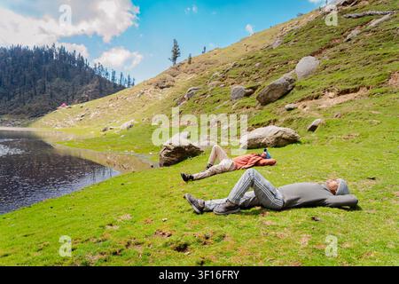 Zwei Wanderer liegen und entspannen sich am grünen grasbewachsenen Ufer des Kareri-Sees in den Bergen von Himachal Pradesh. Stockfoto