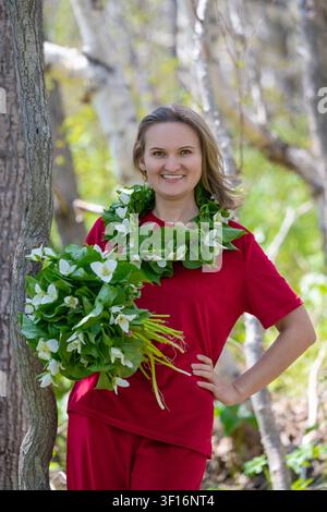 Lächelnde Frau in rotem Outfit steht in sonnigem Wald und hält einen großen Trillienstrauß, während sie einen passenden Blumenkranz trägt. Natürliches Licht hebt das Grün hervor Stockfoto