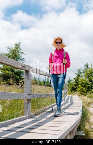 Frau wandert mit Trekkingstöcken auf einem Holzsteg durch Sommerwiese und Nadelwald, Rückansicht mit Rucksack, Hut und Kopffrei unter cl Stockfoto