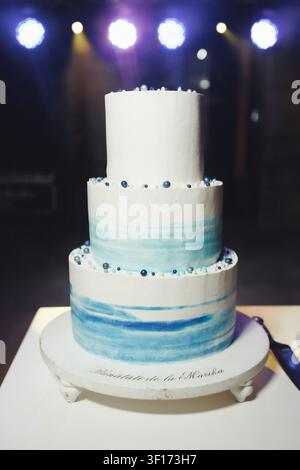 Weißer Kuchen mit blauen Aquarellwirbeln und Perlenakzenten. Dreistöckiges Design steht auf einem eleganten Kuchenständer. Sanfte Bühnenlichter beleuchten das Dessert's Ele Stockfoto