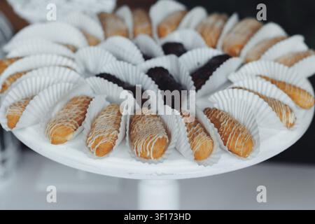 Goldene Gebäckstücke ruhen auf einem weißen Kuchenständer. Jedes Gebäckstück ist in zarte Papierbecher gehüllt. Einige sind mit Zucker bestäubt, andere mit Schokolade beträufelt. T Stockfoto