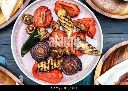 Oberblick auf einen Teller mit verschiedenen gegrillten Paprikaschoten, Tomaten, roten Zwiebeln, Zucchini und Pilzen in der Mitte eines Tisches Stockfoto