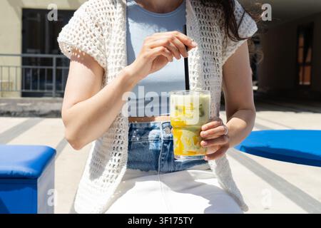 Nahaufnahme einer Frau, die an einem Sommertag in einem Straßencafé sitzt und mit einem Strohhalm einen erfrischenden Mango-Matcha-Latte umrührt Stockfoto