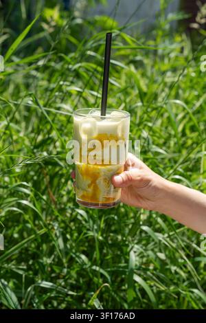 Nahaufnahme einer Frau, die einen eisgekühlten Mango-Matcha-Latte vor üppigem grünem Gras hält Stockfoto