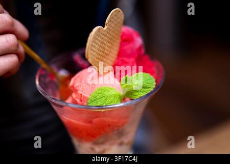 Eine Hand, die einen Löffel hält, schöpft in ein Glas mit rosa Eiscreme. Herzförmiges Waffelstück und Minz-Garnitur. Stockfoto
