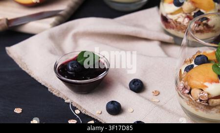 Ein Glasbecher enthält ein geschichtetes Frühstücks-Parfait mit frischen Pfirsichen, Blaubeeren und knuspriger Granola. Sanftes natürliches Licht erhellt die lebendigen Früchte Stockfoto