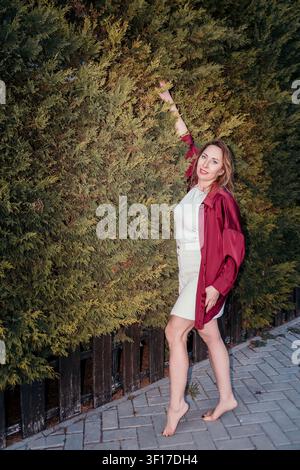 Frau Heckenpark Lächeln, Frau in rotem Hemd, die sich vor einer dichten grünen Hecke in einer Gartenumgebung im Freien posiert Stockfoto