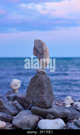 Ausgeglichene Steine, die an einer Meeresküste mit ruhigem Wasser und einem farbenfrohen Sonnenuntergangshimmel angeordnet sind, zeigen die natürlichen Texturen und Formen der Felsen Stockfoto