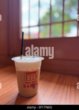 Eiskaffee serviert in einem Plastik-To-Go-Becher mit einem Trinkhalm auf einem Holztisch in einem Café in Bandung, West-Java, Indonesien. Stockfoto