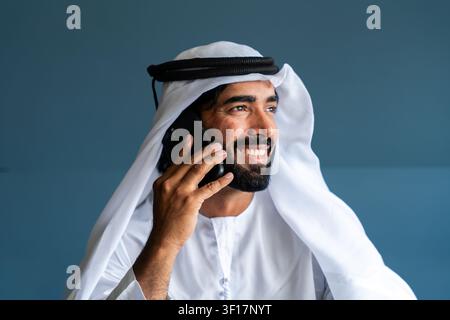Emirati-Mann, der einen Anruf auf seinem Handy führt, lächelnd mit einem glücklichen Gesichtsausdruck, während er jemandem zuhört, zeigt Konzepte von Kommunikation, Technik Stockfoto