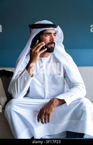 Emirati-Mann in traditioneller Kandura und Ghutra, sitzt auf einem Sofa und führt ein ernstes Geschäftsgespräch an seinem Mobiltelefon und schaut weg Stockfoto