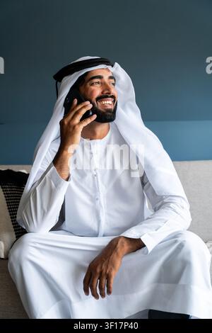 Emirati-Mann mit Bart, der traditionelle Kandura und Ghutra trägt, sitzt auf einem Sofa und telefoniert, drückt Glück und gute Kommunikation aus Stockfoto