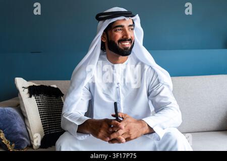 Emiratier mit Bart, der traditionelle Kandura und Ghutra trägt, sitzt auf einem Sofa und schaut mit einem echten Lächeln weg, das Glück und Erfolg widerspiegelt Stockfoto