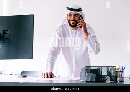Emiratischer Geschäftsmann in traditioneller Kleidung, Ghutra und Kandura, der an seinem Bürotisch steht und ein Gespräch an seinem Mobiltelefon führt, während er auf die Toilette geht Stockfoto