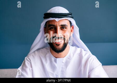Emiratischer Mann mit Bart, der aufrichtig lächelt, den Betrachter direkt ansieht, traditionelle weiße Kandura und Ghutra mit Agal trägt, repräsentiert mi Stockfoto