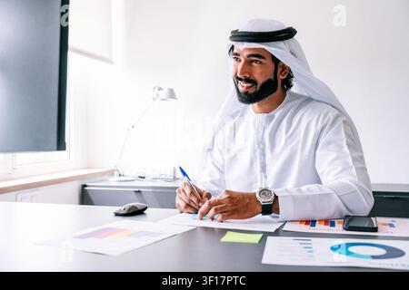 Arabischer Geschäftsmann, der traditionelle Emirati-Kandura und Ghutra trägt, lächelt, während er Papierkram erledigt und Finanzdaten in seinem modernen Büro analysiert Stockfoto