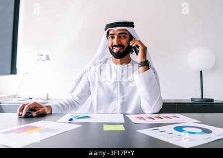 Lächelnder arabischer Mann, der eine traditionelle Kandura und Ghutra trägt, telefoniert und eine Computermaus an seinem Schreibtisch mit Geschäftsgrafiken benutzt, repräsentiert Stockfoto