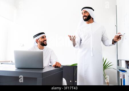 Zwei arabische männliche Geschäftspartner, die traditionelle emiratische Kandura tragen, diskutieren aktiv über eine neue Strategie und lächeln, während sie Ideen auf einem Whiteboard präsentieren Stockfoto
