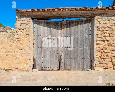 Ein großes, rustikales Holztor mit metallenen Nieten, das in eine alte Stein- und Mörtelwand unter einem strahlend blauen Himmel eingelassen ist, Torralba de los Sisones, Spanien Stockfoto